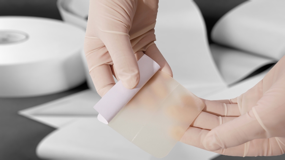 Hands in surgical gloves removing the liner from a hydrocolloid bandage. The out-of-focus background is of different rolls of hydrocolloid tapes.